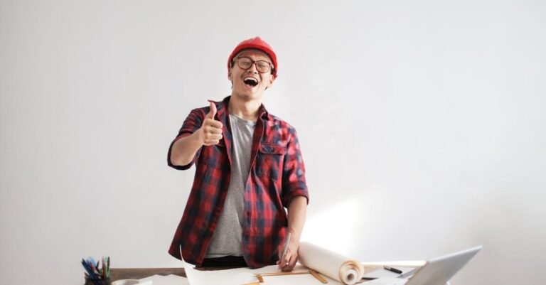 Cheerful male architect in a red hardhat and plaid shirt giving thumbs up at his desk.